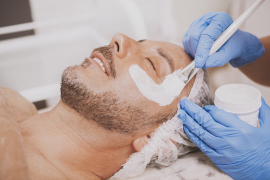 Mature bearded joyful man receiving facial spa treatment, beautician applying mask on the face of male client. Happy man relaxing at spa center, getting face skincare therapy