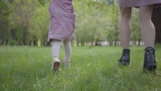 Legs of young mother and little girl running in the park on the green grass holding hands close up. Active healthy lifestyle, connection with nature, carefree childhood