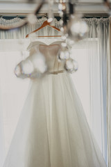 Wedding dress. White wedding dress with a full skirt on a hanger in the room of the bride with white curtains. Wedding attributes. No people.
