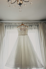 Wedding dress. White wedding dress with a full skirt on a hanger in the room of the bride with white curtains. Wedding attributes. No people.