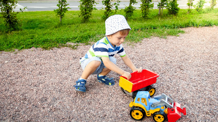 Portrait of cute 3 years old toddler boy sitting on the playground at park and playing with colorful plastic toy truck. Child having fun and playing outdoors with toys