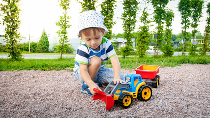 Closeup image of cute little boy playing on the palyground with toys. Child having fun with truck, excavator and trailer. He is pretending to be a builder or driver