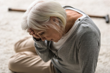 senior woman with migraine sitting on carpet and touching forehead with hand