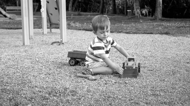 Black and white image of 3 years old toddler boy sitting in the sandbox at palyground and playing with toy truck excavator and trailer