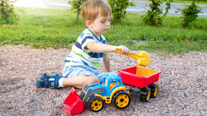 Portrait of cute 3 years old toddler boy sitting on the playground at park and playing with colorful plastic toy truck. Child having fun and playing outdoors with toys