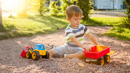 Closeup image of cute little boy playing on the palyground with toys. Child having fun with truck, excavator and trailer. He is pretending to be a builder or driver