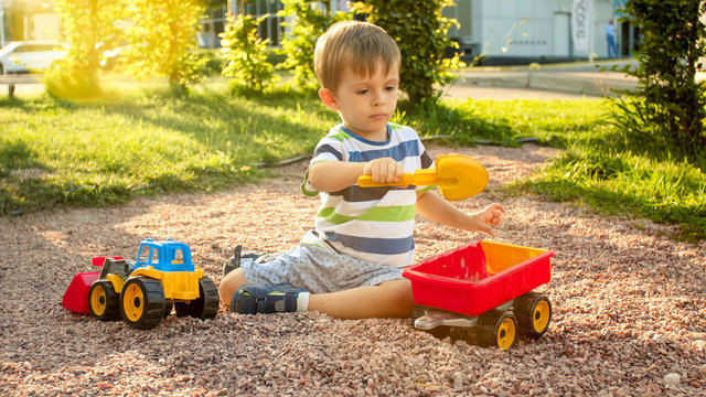Closeup image of cute little boy playing on the palyground with toys. Child having fun with truck, excavator and trailer. He is pretending to be a builder or driver