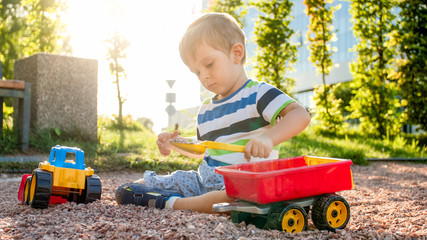 Closeup portrait of happy smiling 3 years old child boy digging sand on the playground with toy plastic truck or excavator. Child palying and having fun at park at summer