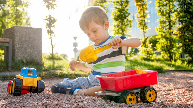 Photo of adorable 3 years old toddler boy playing with sand and you truck and trailer in park. Child digging and building in sandpit