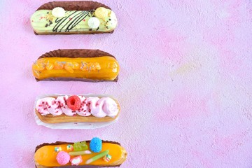Set of design delicious eclairs with creative colorful decor on the pink textured table. Tasty French eclairs with icing, cream, fresh berries and sugar elements. Sweet flat lay. Homemade profiteroles