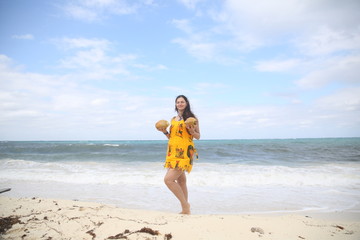Summer holidays and vacations - an attractive girl on the sandy beach of the sea with coconuts in her hands in a yellow dress against a blue sea and sky