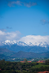 Obraz premium Vertical landscape of valley with houses and with the Picos de Europa snowed in the background in Cantabria, Spain, Europe