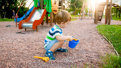 Image of little boy sitting on the playground and puring sand with small plastic spade in colorful bucket. Kid digging and building at park