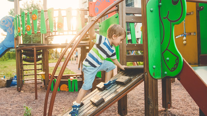 Photo of adorable toddler boy climbing and crawling on wooden staircase on children palyground at...