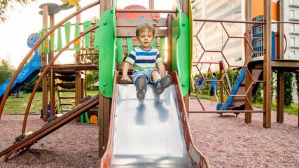 Photo of cute 3 years old toddler boy climbing and riding on big slide on children playground at...
