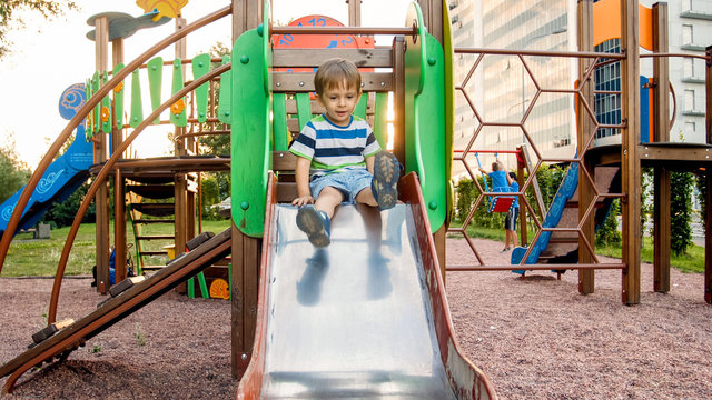 Photo of adorable smiling boy climbing and riding on slide. Active child having fun and playing at park