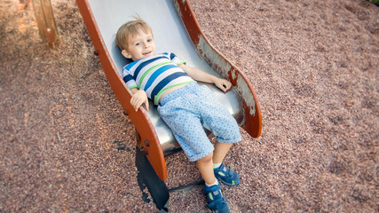 Photo of cute 3 years old toddler boy climbing and riding on big slide on children playground at park