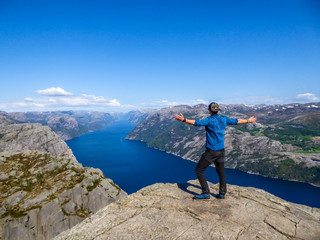 A man wearing blue shirt stands at the edge of a steep cliff of Preikestolen. A view on Lysefjorden. Man enjoys the view, feels free and happy. Man is spreading the arms wide open. Gesture of freedom