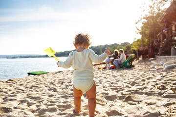 A small child stands on the beach with a toy in his hands. The baby will soon be swimming in the sea.