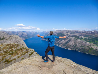 A man wearing blue shirt stands at the edge of a steep cliff of Preikestolen. A view on Lysefjorden. Man enjoys the view, feels free and happy. Man is spreading the arms wide open. Gesture of freedom