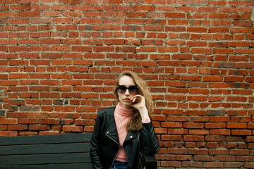 Fototapeta na wymiar blonde girl with curly hair in black glasses, pink turtleneck, in a black leather rocker jacket on the background of a red brick wall, with a place for the inscription on top