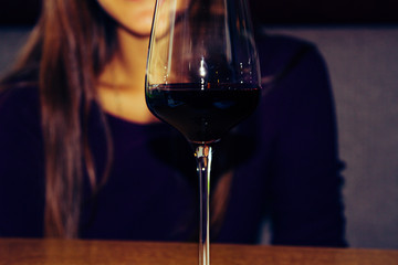 A young girl is sitting in a cafe. Before her is a glass of red wine. The general background of the picture is crimson.