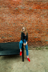 full-length blonde girl with curly hair in black glasses, pink turtleneck, blue jeans, red sneakers in black leather rocker jacket on a red brick wall background,with plenty of space for lettering