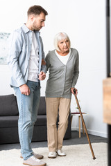 full length view of man helping senior mother with cane at home