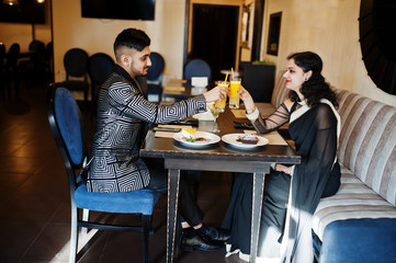 Lovely indian couple in love, wear at saree and elegant suit, sitting on restaurant and cheers together by orange juice.