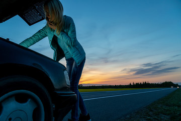 Beautiful slim blonde girl talking on the phone and looking under the hood of a car. The girl will...