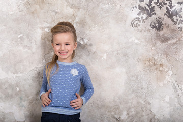 Portrait of a little attractive smiling girl in a blue sweater and pants with hair folded into her hair against a grunge wall background.