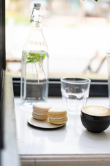 In summer cafe on window tree ficus. Near a bottle of fresh cold water with mint leaves. In a cup of hot cappuccino coffee and on a saucer sweet dessert macarons of light yellow color of pastel tones.