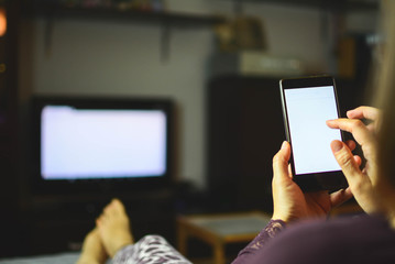 A woman with outstretched legs on the couch in the living room watches the TV and uses a DLNA on a smartphone, wireless internet, social networking. Gambling, mobile betting. Addictions