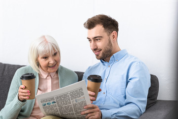 Fototapeta na wymiar smiling man and surprised senior mother holding coffee and reading newspaper at home