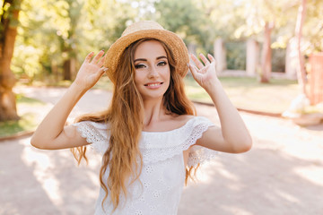 Lovely young lady in trendy white dress walking in park in sunny morning and smiling. Outdoor portrait of pretty fashionable girl posing with pleasure in straw hat.