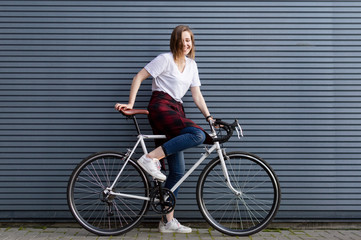 Fototapeta na wymiar beautiful young girl sit down on a white bicycle on the background of a gray striped wall, the woman is happy