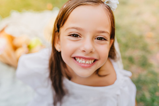 Close-up portrait of dark-eyed girl with cheerful smile posing with pleasure outdoor. Pretty little lady with brown eyes and long eyelashes laughing on blur nature background.