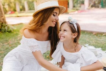 Gorgeous young woman in trendy hat with white ribbon going to kiss daughter in forehead. Laughing...