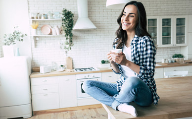 Cute lovely young woman in casual wear with coffee cup in hands is sitting on table in kitchen and relaxing.