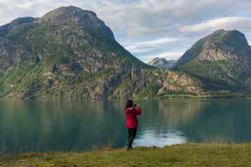 Young caucasian woman in a red jacket   taking a photo with her smartphone in a beautiful scene, on the shore of a lake, with a view towards mountains