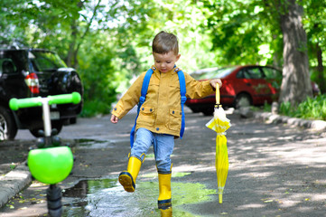  A little cute boy in a yellow raincoat walks through the puddles smiling and holding an umbrella with a lemon coloring.
