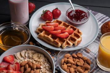 Breakfast. Healthy food - milk oatmeal with a belgian waffle with fruit, honey, strawberry jam, chocolate muffin and fresh yogurt. Set meal on black table background. Top view