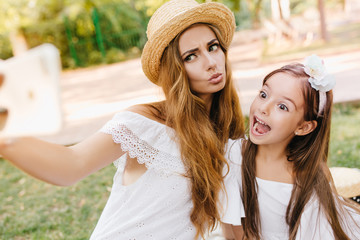 Attractive young woman in lace dress fooling around with daughter for photo, while resting in park. Stylish lady and cute little girl making funny faces for selfie.