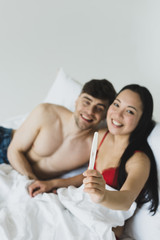 cheerful asian woman looking at camera while holding pregnancy test near smiling boyfriend