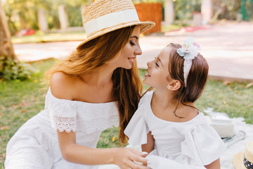 Excited brown-haired girl with ribbon looking at smiling mother with stylish make-up. Outdoor...