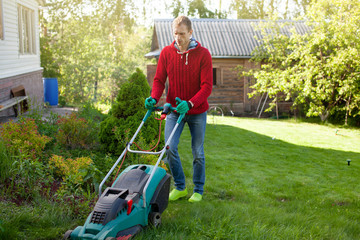 Young man mowing the lawn. Worker doing his job in backyard. Spending summer day in garden. Lawnmower standing on the background of private    wooden house cottage.