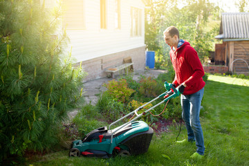 Young man mowing the lawn. Worker doing his job in backyard. Spending summer day in garden. Lawnmower standing on the background of private    wooden house cottage.