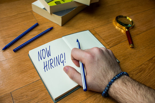 Word writing text Now Hiring. Business photo showcasing finding evaluating working relationship with future employees Close up view man writing notebook pen set lying wooden table two books