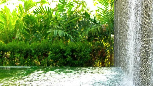 Garden water feature with water falling over a wall covered in small stones into a peaceful pool. Tropical plants and bright warm sunlight in the background. Man made waterfall at a resort hotel.
