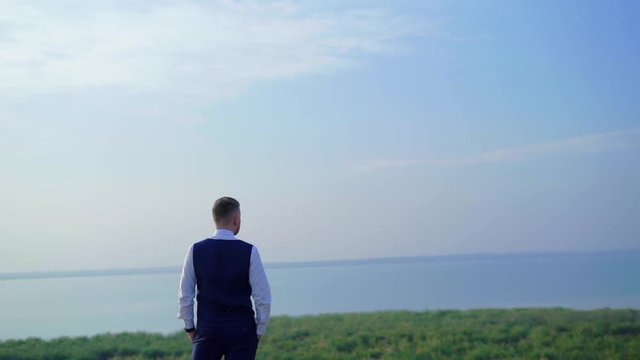Young groom standing near the hill coast with sea on background. Happy wedding moment